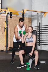 Wall Mural - Smiling woman training with dumbbells near trainer in gym.