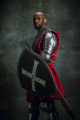 Wall Mural - Medieval knight. Portrait of brutal african man, medieval warrior wearing armour isolated over dark vintage background. Comparison of eras, history