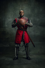 Wall Mural - Vintage style portrait of brutal dark skinned man, medieval warrior or knight wearing armour holding basketball ball isolated over dark background. Comparison of eras, history