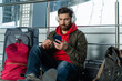 © NFstock - Calm man wearing headphones thoughtfully looking into the smartphone screen while sitting near his luggage and waiting for his flight. Trip concept