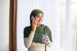 © Prostock-studio - Young Arab woman in hijab standing near window with aromatic coffee, speaking on cellphone at home