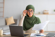 © Prostock-studio - Young Arab female freelancer in hijab working with papers and laptop at home