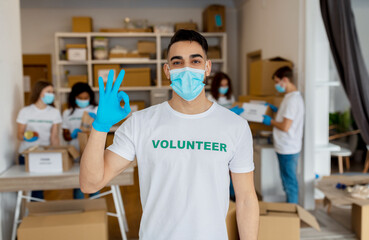 Wall Mural - Volunteering concept. Young arab man in medical mask and gloves showing OK gesture, working in charity center