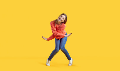 Wall Mural - Funny carefree beautiful young woman having fun. Happy pretty teenage girl in comfortable casual wear looking at camera and smiling while dancing in the studio against vibrant yellow colour background