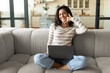 © Prostock-studio - Cheerful young woman doing phone gesture, saying call me back, using laptop, sitting on couch at home, copy space