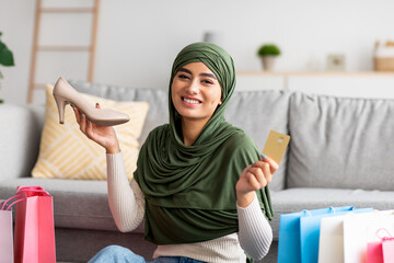 Wall Mural - Young Arab woman in hijab showing credit card and shoe, sitting on floor among gift bags, buying footwear on internet