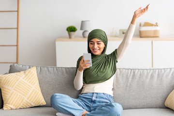 Wall Mural - Young Arab woman playing online game on smartphone, making victory gesture, using new gaming app on mobile device