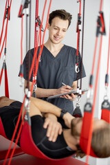 Wall Mural - Rehabilitation specialist examining male patient before active treatment on suspension straps. Therapeutic exercises and neuromuscular activation on red cord slings