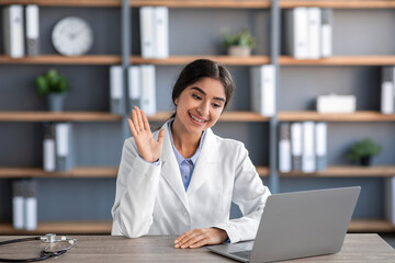 Wall Mural - Friendly cheerful young indian female doctor in white coat works at workplace and waves hand to laptop in clinic