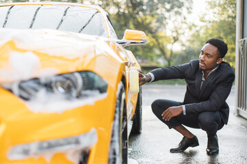 Poster - Luxury modern sport car in soap foam outdoors at car wash service. Side view of handsome young busy african man in suit using yellow sponge for cleaning car door with soap foam outdoors.