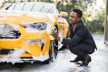 Wall Mural - Car washing outdoors ar self wash service station. Handsome African Confident male entrepreneur in cin business suit, washing his modern sport car headlights with yellow sponge and soap.