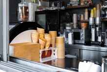 Many Empty Baked Waffle Cones And Paper Cups For Ice Cream Stacked At Display Counter Window Of Street Food Takeaway Cafe Window. Sweet Desserts And Hot Tea Coffee Cafeteria