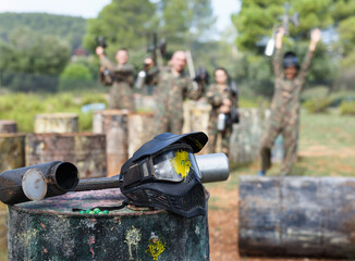 Sticker - View of protective mask with splash of paint and marker gun on blurred background with people in camouflage