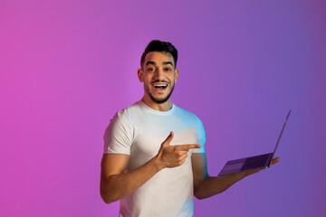 Wall Mural - Cheery young Arab man pointing at laptop computer, working online, communicating on web in neon light