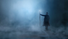 Magician In Cloak, Cowl With Magic Stick Standing In Fog Landscape Illuminated By Blue Moon Ligh. Fantasy, Wizard Concept, 3D Rendering