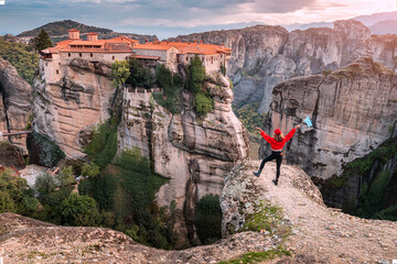 Wall Mural - Happy traveler girl with a Greek flag stands on top of a cliff and admires the grandiose view of the famous Meteora monasteries. Wonders of the world and tourist attractions