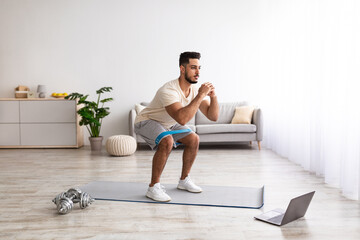 Wall Mural - Full length of young Arab guy working out in front of laptop computer at home, following online video tutorial indoors
