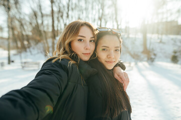 Portrait of two beautiful women in sunny winter forest taking selfie and looking at camera with smile on faces on sunset background.