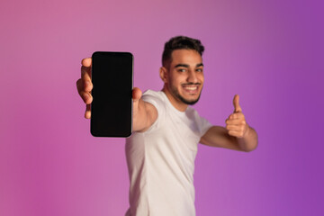 Wall Mural - Cheerful millennial Arab guy pointing at smartphone with blank screen, recommending mobile app in neon light, mockup