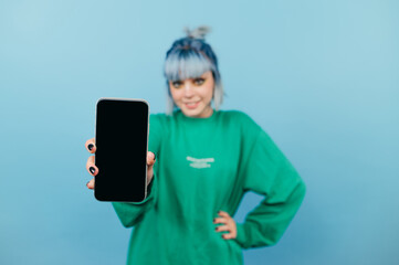 Positive woman in a green sweatshirt stands with a smartphone in his hands on a blue background and shows a black screen to the camera with a smile on his face. Copy space