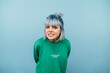 © bodnarphoto - Cute girl with blue hair clenching her teeth looks into the camera and squints her eyes, isolated on a blue background, wearing a green sweatshirt.