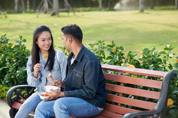 Cheerful young couple sitting on bench in park, talking and eating delicious treet food