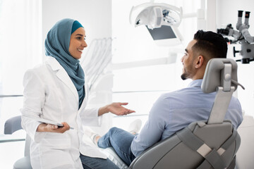 Wall Mural - Muslim Dentist Woman With Digital Tablet Consulting Male Patient About Teeth Treatment