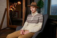 Portrait Handsome Fashionable Man In A White Sweater And Brown Hat Sits On A Chair In A Photo Studio Loft. Young Businessman Is Thinking About Future