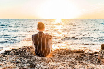 Wall Mural - A young blond man stands on the seashore in front of a bright orange sunset. Romantic seascape