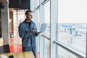 Portrait of a smart intelligent male manager, he is holding a laptop for research in the office, a business man in elegant clothes playing on the touchpad. Standing in the office by a large window