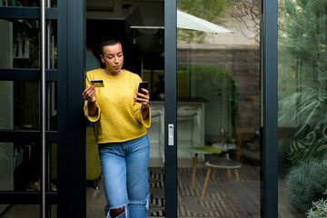 Wall Mural - Portrait of casual relaxed woman using phone for online payment on doorstep of her home shopping online from home leaning on door frame using credit card casual entrepreneur doing business from home