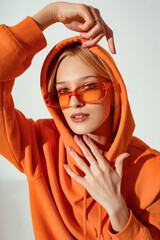 Wall Mural - Fashion portrait of confident woman wearing trendy orange color sunglasses, oversized hoodie. Model looks at camera. Indoor, studio close up fashion portrait