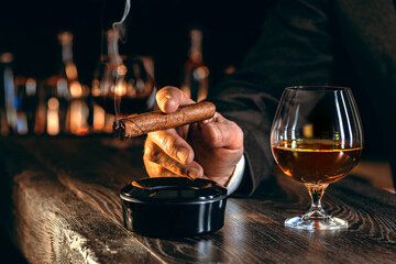 Man's hands with a cigar, elegant glass of brandy on the bar counter. Alcoholic drinks, cognac, whiskey, port, brandy, rum, scotch, bourbon. Vintage wooden table in a pub at night