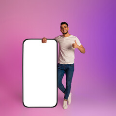 Wall Mural - Millennial Arab man standing near giant smartphone with empty white screen, gesturing thumb up in neon light, mockup