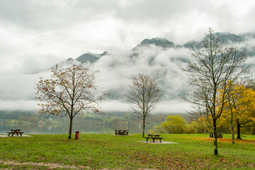 Moody autumn landscape with bare trees and benches on mountain lake shore.