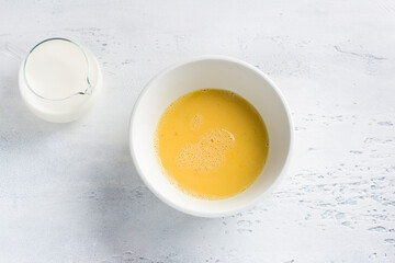 A white bowl with beaten egg and jug of milk on light blue background, top view. Cooking omelet, homemade healthy food