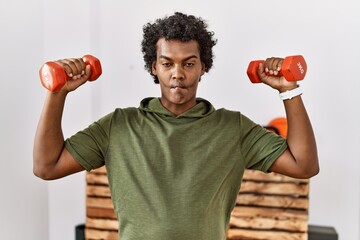 Poster - African man with curly hair wearing sportswear using dumbbells making fish face with mouth and squinting eyes, crazy and comical.