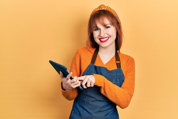 Poster - Redhead young woman wearing waitress apron holding touchpad device taking order looking positive and happy standing and smiling with a confident smile showing teeth