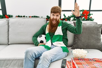 Sticker - Redhead man with long beard football hooligan holding ball sitting on the sofa celebrating victory with happy smile and winner expression with raised hands