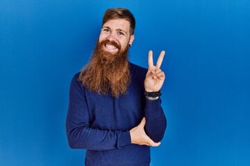 Wall Mural - Redhead man with long beard wearing casual blue sweater over blue background smiling with happy face winking at the camera doing victory sign with fingers. number two.