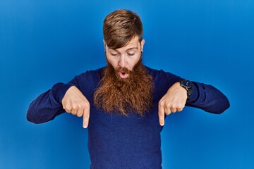 Canvas Print - Redhead man with long beard wearing casual blue sweater over blue background pointing down with fingers showing advertisement, surprised face and open mouth