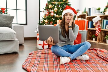 Sticker - Young latin woman using laptop sitting by christmas tree afraid and shocked, surprise and amazed expression with hands on face