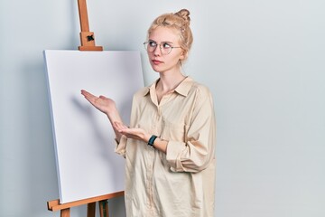 Wall Mural - Beautiful caucasian woman with blond hair standing by painter easel stand presenting canva smiling looking to the side and staring away thinking.