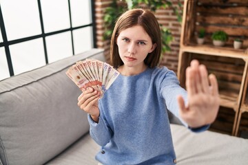 Sticker - Young brunette woman holding colombian pesos with open hand doing stop sign with serious and confident expression, defense gesture