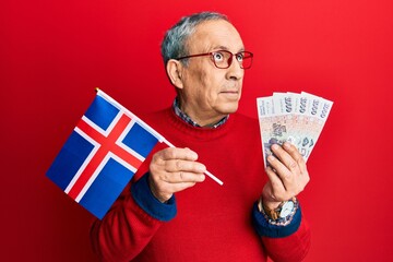 Canvas Print - Handsome senior man with grey hair holding iceland flag and icelandic krona banknotes smiling looking to the side and staring away thinking.