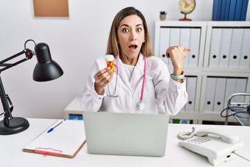 Canvas Print - Young hispanic woman wearing doctor uniform holding pills at the clinic surprised pointing with hand finger to the side, open mouth amazed expression.