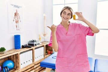 Poster - Young physiotherapist woman working at pain recovery clinic holding pills pointing thumb up to the side smiling happy with open mouth