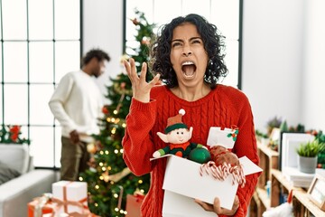 Poster - Young hispanic woman standing by christmas tree with decoration crazy and mad shouting and yelling with aggressive expression and arms raised. frustration concept.