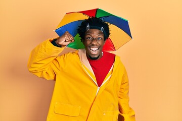 Sticker - Young african american man wearing yellow raincoat angry and mad raising fist frustrated and furious while shouting with anger. rage and aggressive concept.