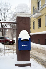 Hanging From A Fence Post Is A Blue Mailbox With A Snow Cap On The Roof. In The Background Is A House And A Car Leaving The Yard.
